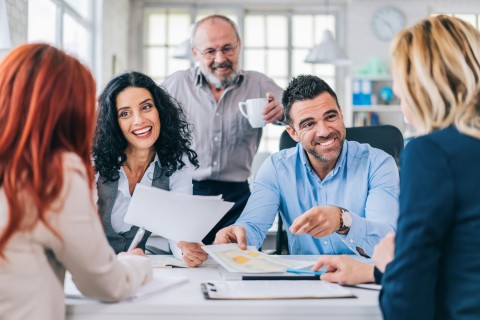 Mehrere Personen um einen Tisch mit Unterlagen und Warmgetränken, diskutieren in enspannter Stimmung